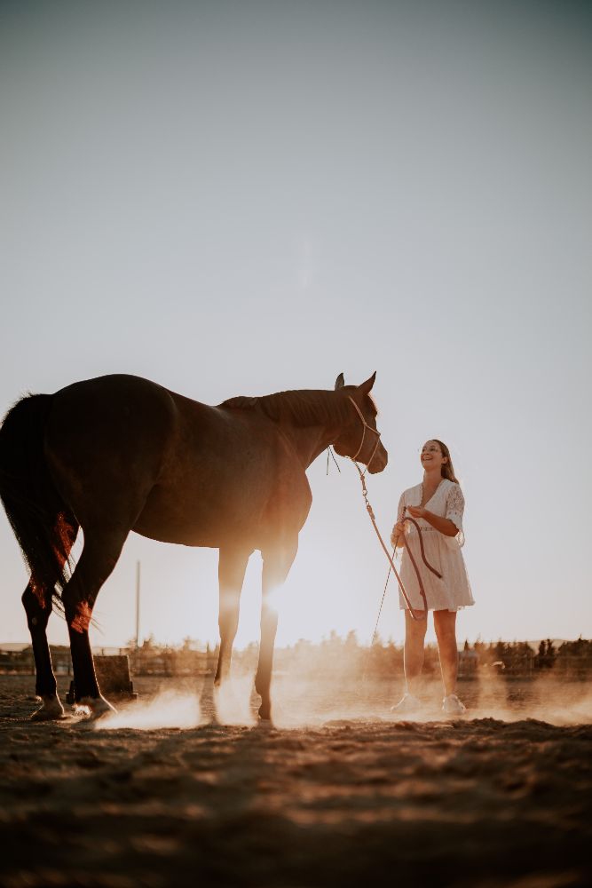 Une femme en robe blanche tient son cheval à contre-jour
